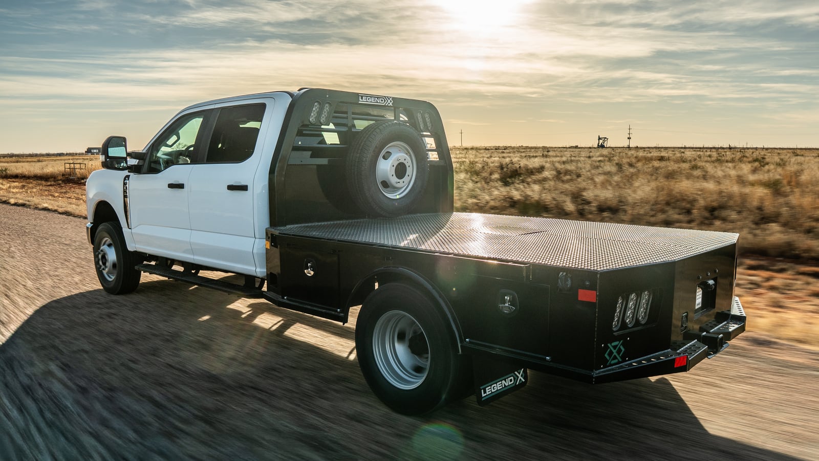White pickup with a black Legend X Viking flatbed driving on a rural road at golden hour.